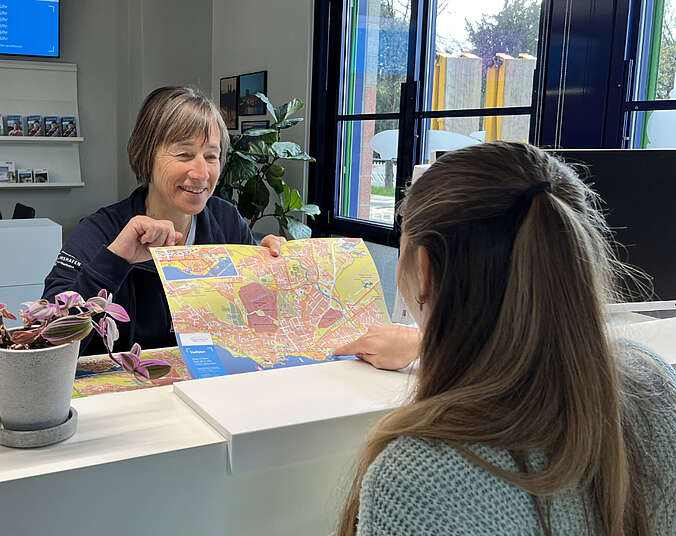 Frau erklärt hinter Tresen anderer Frau Weg anhand Stadtkarte